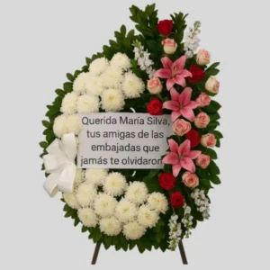 Corona asimétrica floral con lirios rosados, rosas rojas y crisantemos blancos, diseño vertical montado en trípode con cinta blanca personalizada y moño satinado. Arreglo fúnebre elegante para condolencias.