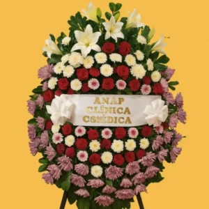 Corona colorida fúnebre con lirios blancos, rosas rojas y crisantemos púrpura, diseño ovalado montado en soporte con cinta blanca personalizada y moños satinados. Arreglo floral solemne para condolencias.