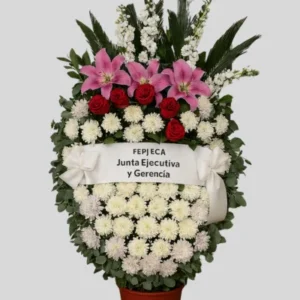 Corona en maceta con lirios rosados, rosas rojas y crisantemos blancos, diseño ovalado con cinta blanca personalizada y moño satinado. Arreglo fúnebre robusto y elegante.