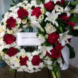 Corona fúnebre con rosas rojas, lirios blancos, crisantemos y rosas crema, diseño circular montado en trípode con cinta blanca personalizada. Arreglo floral elegante para condolencias.