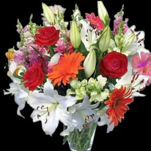 Ramo de flores vibrante con rosas rojas, lirios blancos y gerberas naranjas en jarrón de cristal sobre fondo oscuro.