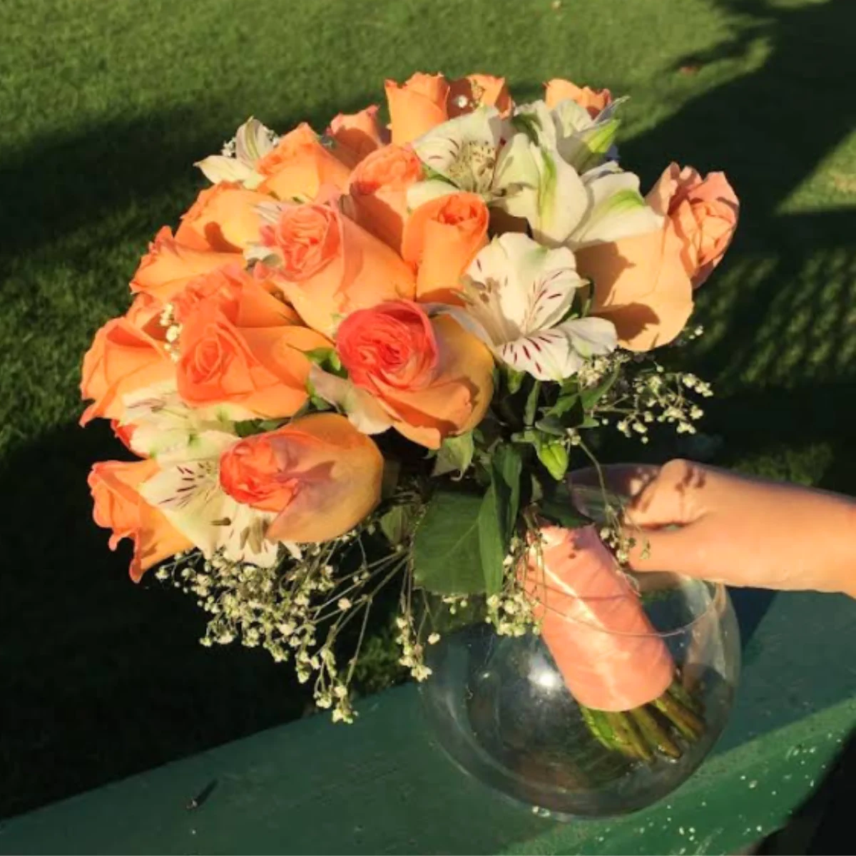 Centro de mesa con rosas color durazno, lirios blancos con vetas rosadas, flores blancas pequeñas y follaje verde en jarrón de vidrio. Ideal para celebraciones y decoración elegante.