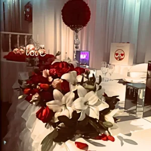 Mesa decorada con rosas rojas, lirios blancos, cristalería, cortinas iluminadas y elementos románticos. Ideal para bodas y celebraciones de amor.