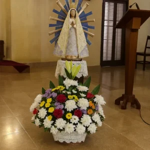 Altar floral mariano con imagen coronada de la Virgen del Rosario sobre pedestal con encaje, rodeada por arreglo multicolor de crisantemos, rosas, margaritas y follaje verde. Composición devocional para celebraciones religiosas.