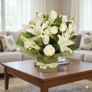 Un arreglo floral de Armonía Blanca Floral con rosas blancas, lirios blancos y otras flores en tonos claros, presentado en una caja cuadrada verde con un lazo, sobre una mesa de madera en un living con sofás claros y alfombra beige.