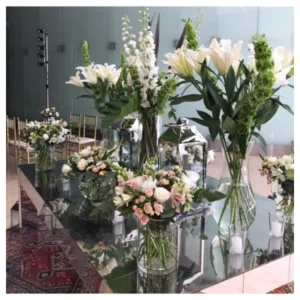 Arreglo floral elegante con lirios blancos, rosas en tonos pastel y follaje verde en jarrones de vidrio sobre mesa espejada, un diseño sofisticado y equilibrado ideal para recepciones, bodas y eventos especiales.