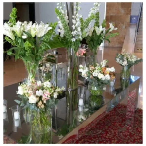 Arreglo floral moderno con lirios blancos, rosas en tonos suaves y flores de temporada en jarrones de vidrio sobre mesa espejada, un diseño fresco y elegante ideal para recepciones, bodas y celebraciones.