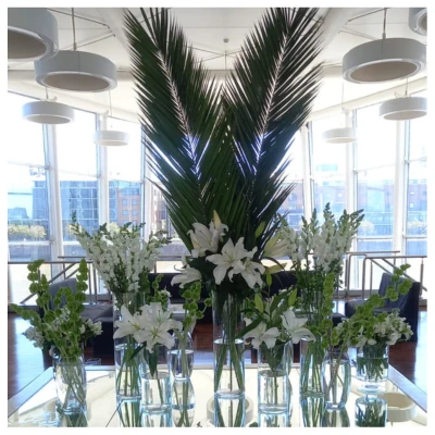 Arreglo floral tropical con lirios blancos, flores de temporada y grandes hojas de palmera en jarrones de vidrio, un diseño moderno, fresco y elegante ideal para recepciones y eventos especiales.