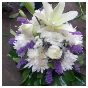 Arreglo floral violeta con lirios blancos, rosas marfil, crisantemos y flores moradas en composición armónica con follaje verde, un diseño elegante y equilibrado ideal para mesas, recepciones y eventos especiales.