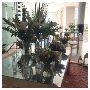 Arreglo mesa elegante con lirios blancos, rosas en tonos suaves y follaje verde en jarrones de vidrio sobre base espejada, un diseño moderno y sofisticado ideal para bodas, recepciones y celebraciones.