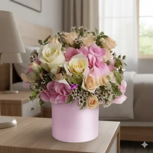 Una Caja Rosas Tonos, un arreglo floral con rosas en tonos pastel de rosa, crema y melocotón, hortensias y follaje, presentado en una caja redonda rosa sobre una mesa de noche de madera clara en un dormitorio.