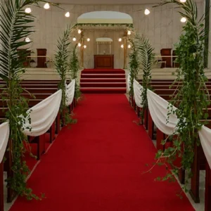 Camino floral ceremonial con alfombra roja, bancos decorados con telas blancas, palmas arqueadas y chuppah blanco con detalles dorados. Montaje escenográfico ideal para bodas religiosas y eventos solemnes.