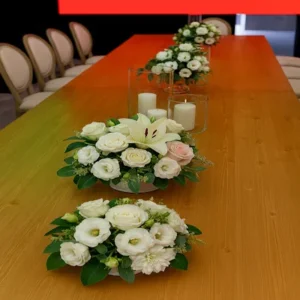 Centro blanco lineal con rosas blancas, lisianthus y follaje verde, intercalado con portavelas de vidrio sobre mesa de madera. Arreglo decorativo ideal para eventos formales y recepciones elegantes.