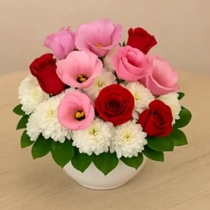 Centro floral compacto con rosas rojas, lisianthus rosados y crisantemos blancos tipo pompon, dispuestos en caja con follaje verde. Arreglo redondo ideal para eventos, regalos o decoración elegante.