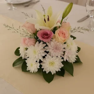 Centro floral elegante con lirios blancos, rosas rosadas y crisantemos sobre base cerámica, ubicado en mesa con mantel crema y vajilla formal. Arreglo decorativo ideal para bodas y eventos.