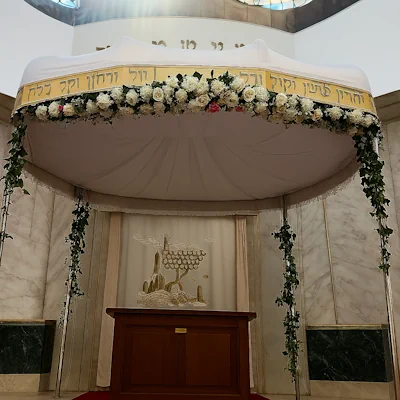 Chuppah floral ceremonial con guirnalda de rosas blancas y rosadas, follaje verde, cubierta blanca con bordes dorados y texto hebreo bordado, enmarcado por fondo de mármol y cortinaje simbólico. Montaje escenográfico para bodas judías.