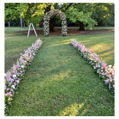 Pasillo floral boda en jardín con sendero de césped enmarcado por arreglos de flores rosadas y blancas que conduce a un arco floral, un diseño romántico y elegante ideal para ceremonias al aire libre.