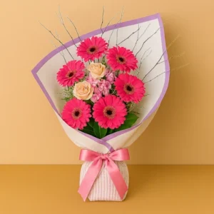 Ramo Gracias Gerberas con flores rosadas, ramas decorativas, envoltura blanca con borde lavanda y lazo texturado, sobre fondo beige y mesa clara. Composición circular, alegre y armoniosa.