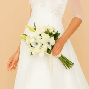 Ramo blanco nupcial con lirios, rosas marfil y follaje verde, sostenido por novia con vestido bordado. Arreglo floral elegante y atemporal ideal para ceremonias de boda.