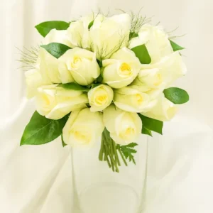 Ramo de rosas blancas redondo, compacto y elegante, adornado con follaje verde y ramitas decorativas, presentado en florero de vidrio transparente. Arreglo ideal para novias y ocasiones especiales.